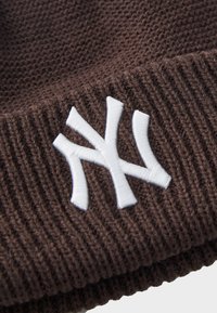 Ruskea neulottu pipo, jossa on ribbireunus ja suuri valkoinen brodeerattu "NY"-logo etupuolella.