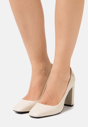 Pies que llevan zapatos de tacón alto beige con punta cuadrada y tacón bloque, contra un fondo blanco liso.