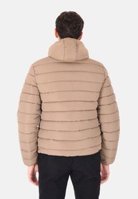 Uomo che indossa un giubbotto imbottito beige con cappuccio, in piedi con la schiena rivolta verso la fotocamera su uno sfondo chiaro e neutro.