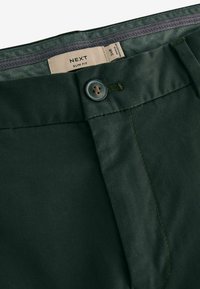 Grüne Slim-Fit-Hosen mit einer glatten Textur, Knopfverschluss und kontrastierenden Nähdetails. Innen ein sichtbares Stoffetikett.