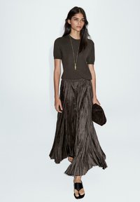 Haut en tricot marron à manches courtes associé à une jupe plissée fluide marron. Le mannequin tient une pochette marron et porte des sandales à talons noires.