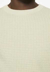 Hellecremefarbene Sweater mit einem strukturierten, gerippten Muster, runder Halslinie und dezenten Nähdetails. Weich gestricktes Material.