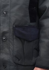 Prim-plan al jachetei negre Barbour, cu butoane și clapetă din catifea pe un braț și o mână vizibile.