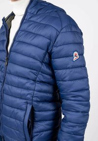 Giacca puffer blu navy con texture trapuntata, zip frontale e tasche laterali. Presenta un patch con il logo sulla manica sinistra.