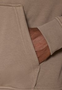 Bežová mikina z látky s ribovým vzorom, so predným vreckom a priliehavými manžetami. Viditeľné švy zvýrazňujú upravený vzhľad.