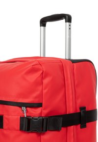 Sac de voyage rouge avec une surface lisse et texturée, doté d'une sangle et d'une fermeture éclair noires, ainsi que d'une poignée rétractable en argent.