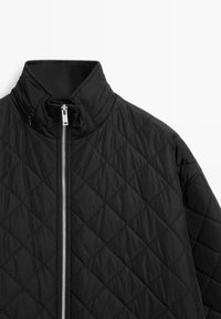 Schwarze gesteppte Jacke mit hohem Kragen und einem langen silbernen Reißverschluss. Der Stoff hat eine glatte, leicht glänzende Textur.