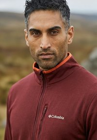 Hombre con cabello corto y rizado que lleva una chaqueta polar Columbia burdeos con cremallera al aire libre, con un fondo natural desenfocado.