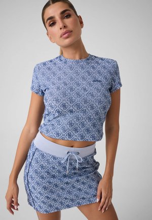 Kvinde med håret bundet tilbage iført matchende blå mønstret kort t-shirt og nederdel med snøre ved taljen mod en ensfarvet baggrund.