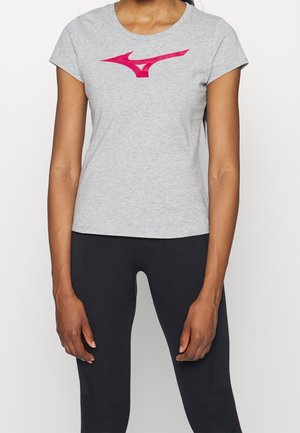 Woman wearing light grey t-shirt with pink logo and black pants, standing against a plain white background.