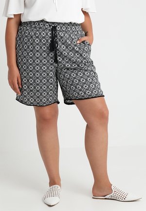 Persoon draagt zwart-witte shorts met bloemenpatroon, een aantrekkoord in de taille en witte geweven instapschoenen tegen een effen achtergrond.