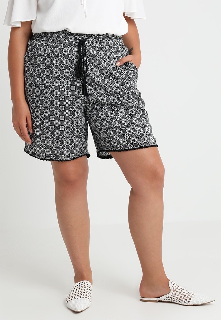Personne portant un short noir et blanc à motifs floraux avec une taille à cordon et des chaussures blanches tissées sans lacets sur un fond uni.