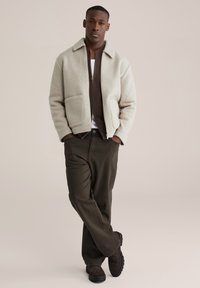 Jachetă crem din amestec de lână cu design cu guler, două buzunare frontale, asortată cu un pulover maro cu fermoar și pantaloni verde închis. Pantofi din piele intoarsă negri.