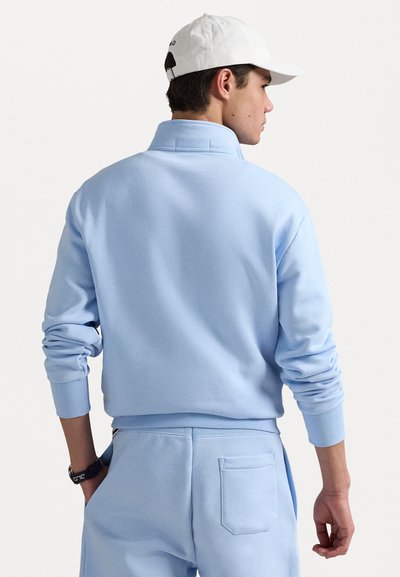 Jasnoniebieski sweter z zamkiem przy kołnierzu, ściągaczami przy mankietach i fasonem typu pullover. Wygląda na miękki, wykonany z teksturowanej tkaniny. Noszony z pasującymi spodniami.