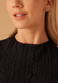 Schwarzes, strukturiertes Oberteil mit hohem Kragen und gewelltem Design, getragen mit einem kleinen silbernen Creolenohrring. Weicher Hautton im Fokus.