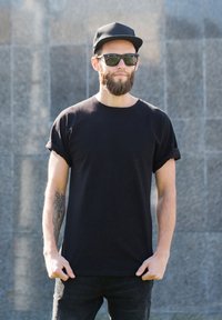T-shirt di cotone nero con maniche arrotolate, indossata con jeans neri. Gli accessori includono un cappellino nero e occhiali da sole scuri. Tatuaggi visibili della tonalità della pelle.
