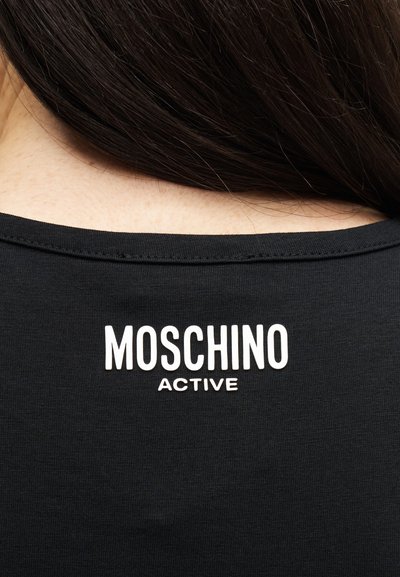 Detailní záběr zadního výstřihu černé košile s nápisem "MOSCHINO ACTIVE" vytištěným bílou barvou pod tmavě hnědými vlasy.
