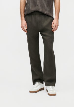 Mężczyzna w ciemnooliwkowych spodniach o prostych nogawkach i białych trampkach, stoi na tle jasnego, jednolitego tła.