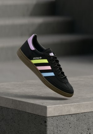 Svart Adidas Spezial-sneaker med pastellgule, rosa og blå striper, lilla hælkappe og brun såle svever over en betongplattform.