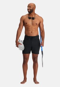Pantaloni da bagno neri con cordino, tenendo una borsa a fantasia in una mano e un boccaglio nell'altra. L'uomo sta in piedi a piedi nudi, sorridente.