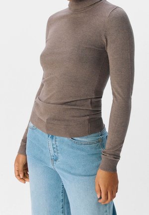 Personne portant un pull à col roulé marron ajusté et un jean taille haute bleu clair sur un fond blanc uni.