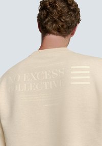 Beige sweatshirt met een gestructureerde stof, geribde ronde hals en een geprinte graphic met de tekst "NO EXCESS COLLECTIVE" op de achterkant.