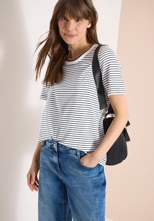 Femme aux longs cheveux bruns portant un t-shirt à manches courtes blanc et noir à rayures, un jean bleu et un sac à bandoulière noir, debout avec une main dans la poche.