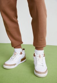 Vita sneakers med bruna accenter, strukturerade sidopaneler och en intrikat mönstrad sula, kombinerat med avsmalnande bruna sweatpants.