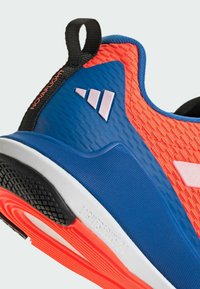 Scarpa sportiva con una tomaia arancione texturizzata, lati blu e un tallone nero. Suola in gomma texturizzata con sottostrato rosa e dettagli di branding.