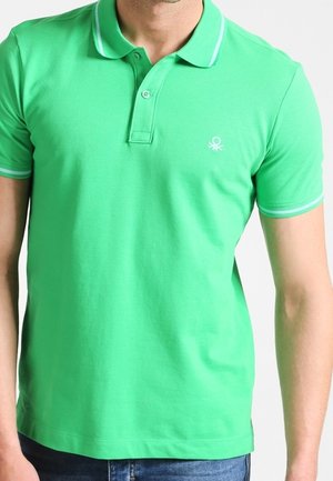 Grünes kurzärmeliges Poloshirt mit weißer Einfassung an Kragen und Ärmeln, getragen von einem männlichen Modell mit blauen Jeans.