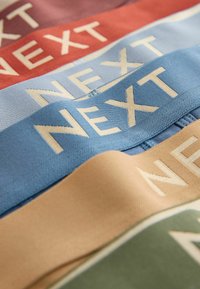 Pinotut pino kankaita punaisena, vaaleansinisenä, sinisenä, beigeenä ja vihreänä, joihin on kudottu suuret vaaleankermanväriset kirjaimet "NEXT".