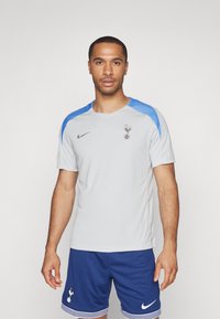 Sivá športová košeľa s modrými akcentmi, obsahujúca logo a branding Nike. Nosená s modrými šortkami so prúžkovaným lemom. Ľahký materiál.