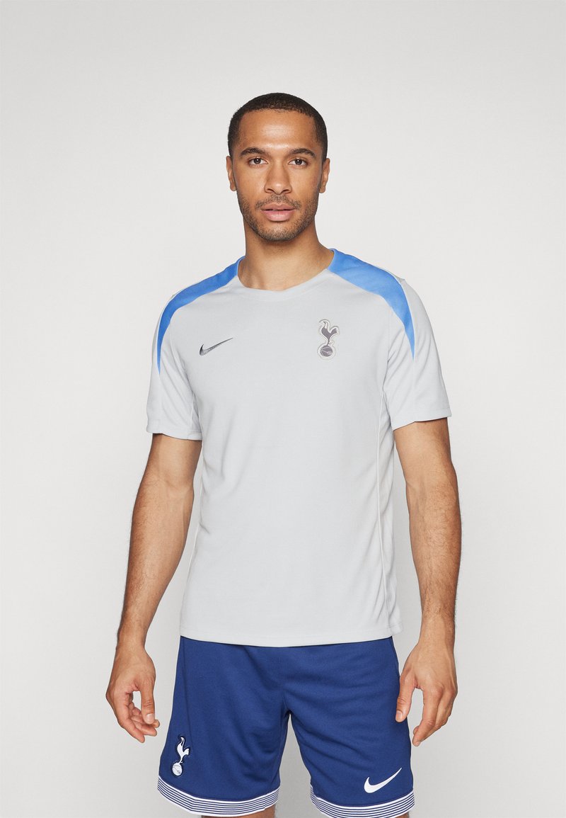 Sivá športová košeľa s modrými akcentmi, obsahujúca logo a branding Nike. Nosená s modrými šortkami so prúžkovaným lemom. Ľahký materiál.