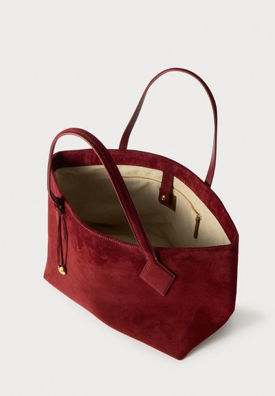 Bordo suede torba s glatkom vanjštinom, otvorom na vrhu, s dva ručka i bež unutrašnjošću koja sadrži zip džep i zlatne metalne detalje.