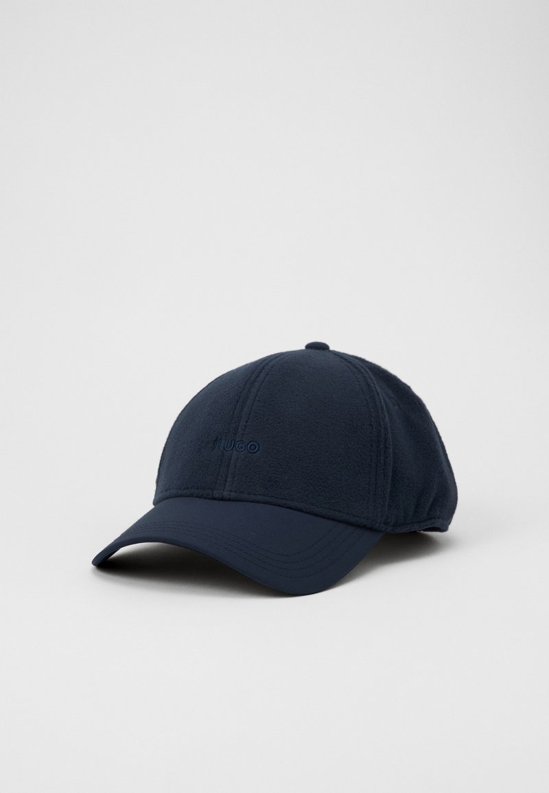 Casquette de baseball bleu marine avec une visière incurvée et un logo "HUGO" discrètement brodé à l'avant, posée sur un fond uni gris clair.