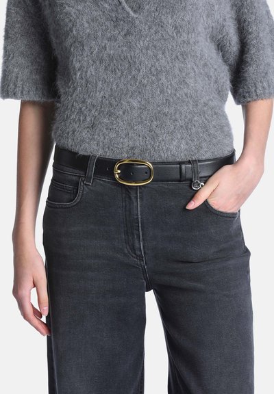 Personne portant un pull gris texturé à manches courtes, un jean noir et une ceinture en cuir noir avec une boucle ovale dorée, main dans la poche.