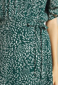 Grön klänning med ett vitt lövmönster, lättviktigt tyg, elastisk midja och knytdetalj vid midjan.
