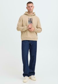 Felpa beige con stampa grafica frontale, abbinata a jeans in denim blu scuro e sneakers crema, in piedi contro uno sfondo chiaro.