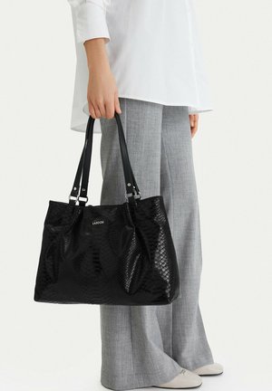 Žena držící velkou černou texturovanou kabelku s dvojitými uchy, oblečená v šedých kalhotách a bílém tričku, stojící na bílém pozadí.