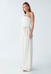 Bela rebrasta majica z enim ramenom, kombinirana z bež hlačami širokih nogavic visoko v pasu z vezanjem spredaj. Gladka tekstura in ohlapen kroj.