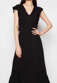 Musta, polvipituinen mekko V-pääntiellä, leppoisilla hihoilla, pleidetulla vartalolla ja kerrotulla hameella, jossa on sivuhalkio. Sileä kankaan tekstuuri.