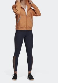 Sweat à capuche orange zippé en tissu doux, avec des accents de zip argentés. Associé à un legging noir avec des panneaux en mesh et des baskets légères.