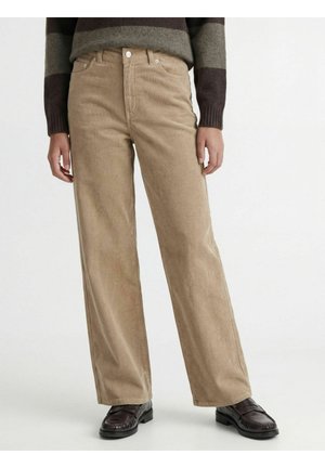 Personne portant un pantalon large en velours côtelé beige, des mocassins marron et un pull rayé brun foncé et gris sur un fond blanc uni.
