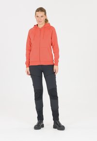 Koraalkleurige rits-hoodie met een buidelzak, gecombineerd met zwarte broeken met ritszakken en een gestructureerde stof. Zwarte laarzen maken de outfit compleet.