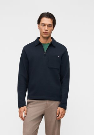 Młody mężczyzna noszący granatowy pulower z suwakiem do połowy i kieszenią na piersi oraz beżowe spodnie, stojący na tle jednolitego jasnego tła.