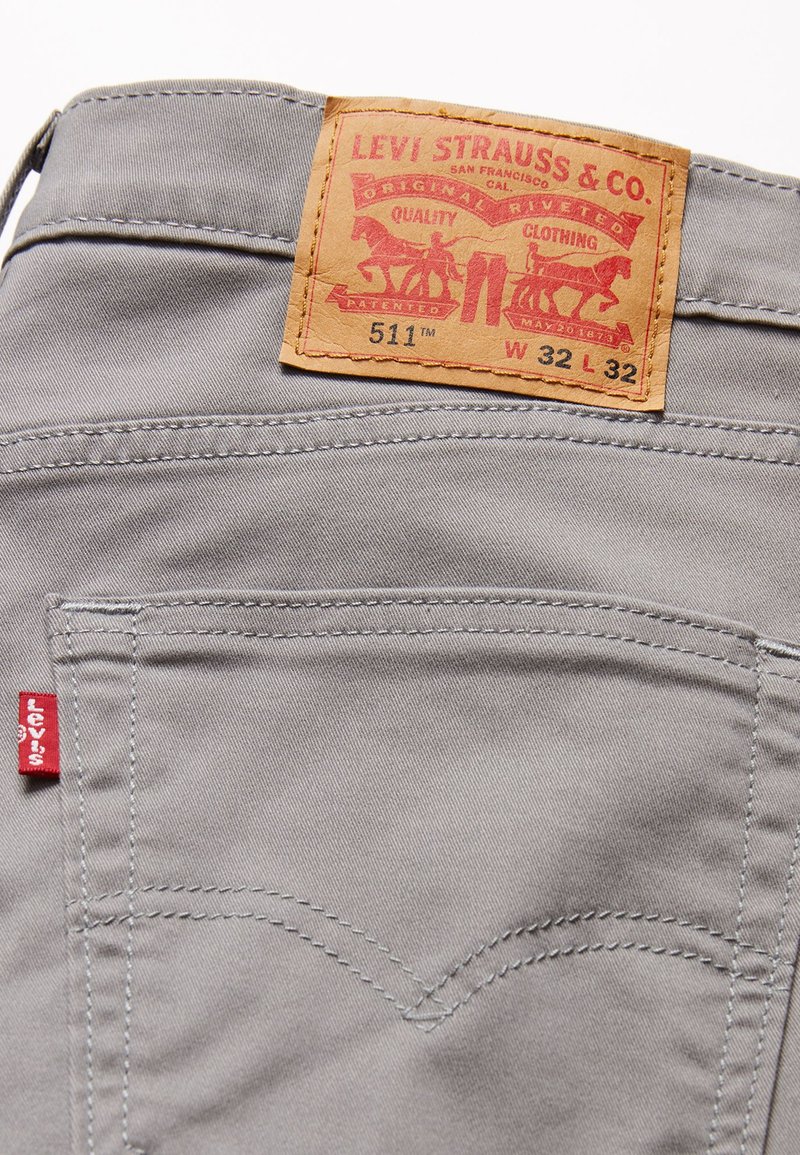 Jean gris avec une étiquette en cuir arborant un texte rouge et jaune, des coutures classiques sur les poches arrière, et un petit onglet rouge sur le côté.