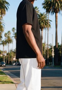Homme portant un t-shirt noir et un pantalon blanc, debout dans une rue ensoleillée bordée de palmiers et de voitures garées.