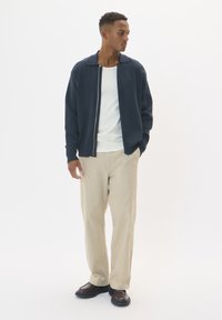 Marinblå zip-up jacka över en vit ribbad skjorta, kombinerad med beigefärgade vida byxor och mörkbruna skor. Slät tyg med en öppen krage design.