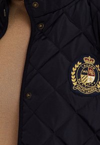 Tmavě modrá prošívaná bunda se zlatým vyšívaným logem Ralph Lauren na levé části hrudi. Má patentní knoflíky a hladkou strukturu.