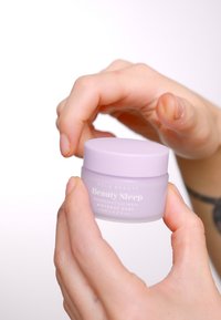 Lilla krukke med et hvit lokk merket "NCLA Beauty Beauty Sleep" for en nattleppe maske, holdt av en hånd med en tatovering.